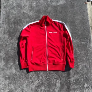 Röd Palm Angels Tracksuit  - Säljer en snygg röd tracksuit från Palm Angels med vita detaljer i utmärkt skick. Jackan har en dragkedja framtill och vita ränder längs ärmarna. Riktigt cool, priset är förhandlingsbart. Köpt från Harrods i London. 