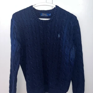Mörkblå stickad tröja från Polo Ralph Lauren - Säljer en snygg mörkblå stickad tröja från Polo Ralph Lauren. Tröjan har ett klassiskt flätmönster och en liten broderad logga på bröstet. Perfekt för kyligare kvällar under vår och sommar! Passar S/M. Sitter bra på någon runt 178-182