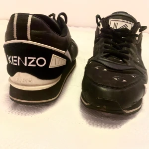 Svarta sneakers från Kenzo - Snygga svarta sneakers från Kenzo med unikt ögonmönster. Skorna har snörning och en rund tå. Perfekta för dig som vill ha en stilren men ändå iögonfallande look. Kenzo-loggan syns tydligt på hälen och sulan.
