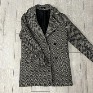 Mönstrad kappa från Oscar Jacobson - Snygg kappa från Oscar Jacobson med ett klassiskt fiskbensmönster i svart och vitt. Den har långa ärmar och knäpps med knappar framtill. Perfekt för höst och vinter med sin stilrena design.