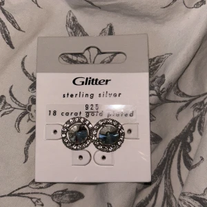 Glittriga örhängen från Glitter - Snygga örhängen från Glitter i sterling silver med 18 karat guldplätering. De har en glittrig design med en stor sten i mitten omgiven av mindre stenar. Perfekta för att ge en extra touch till din outfit.