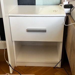 Vit sängbord med låda - Praktiskt vitt sängbord med en låda och en öppen hylla. Perfekt för förvaring av böcker, lampor eller andra småsaker. Stilren design med en enkel handtag i silverfärg. Passar bra i sovrummet för en modern och funktionell inredning. Använt skick. Se bild 3. ENDAST UPPHÄMTNING 