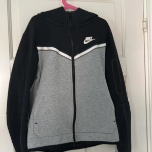 Nike tech ziphoodie i svart och grå - Nike hoodie med dragkedja i svart och grått. Jackan har huva, vita detaljer och logga på bröstet. Praktiska fickor framtill och en extra ficka med dragkedja på ärmen. Perfekt för träning eller chill.