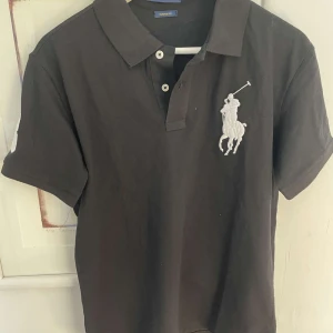 Svart pikétröja från Ralph Lauren - Klassisk svart pikétröja från Ralph Lauren med stor vit broderad logga på bröstet. Perfekt för en stilren och sportig look.