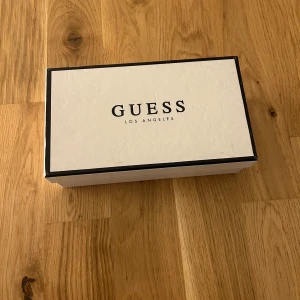 Röd plånbok från Guess - Säljer en snygg röd plånbok från Guess med avtagbar handledsrem och guldfärgade detaljer. Plånboken har flera fack för kort och sedlar samt ett genomskinligt fönster för ID. Klassisk Guess-logga framtill.