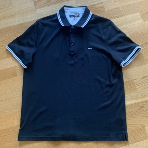 Svart pikétröja från Michael Kors - Säljer en svart pikétröja från Michael Kors med vit logga på bröstet. Tröjan har klassisk krage med vita ränder och knäppning med tre knappar. Ärmarna är korta och har vita ränder vid muddarna. Perfekt för en stilren look. Knappt använd och perfekt inför sommaren. Pris på nätet 1300 kr. 