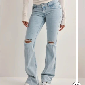 Ljusblå bootcut jeans med slitningar - Säljer ett par ljusblå jeans med bootcut passform och slitna detaljer vid knäna. Jeansen har klassisk femficksdesign och stängs med knapp och dragkedja. Perfekta för en avslappnad och trendig look.