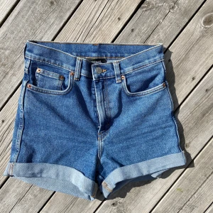 Blå jeansshorts från Monki - Nyskick! Snygga blå jeansshorts från Monki med klassisk femficksdesign och uppvikta benslut. Shortsen har normal passform och hög midja, perfekta för varma dagar. Tidlös modell som är enkel att matcha med olika toppar.