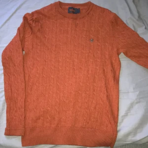 Orange kabelstickad tröja från Morris - Säljer en orange kabelstickad tröja från Morris i 100% ull. Tröjan har rund halsringning, lång ärm och en liten broderad logga på bröstet. Perfekt för dig som gillar klassisk och stilren design.