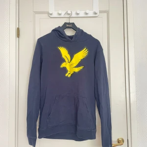Mörkblå hoodie med gul örn från Lyle & Scott - Säljer en mörkblå hoodie från Lyle & Scott med stor gul örn på bröstet. Tröjan har huva och en klassisk magficka framtill. Perfekt för en avslappnad stil och enkel att matcha med jeans eller joggers.