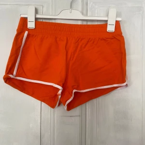Orange shorts med vit kant från H&M - Säljer ett par orange shorts från H&M i 100% bomull. De har elastisk midja och vita kanter längs benen för en sportig look. Perfekta för sommardagar eller träning. Superbekväma och lätta att matcha med en enkel topp.