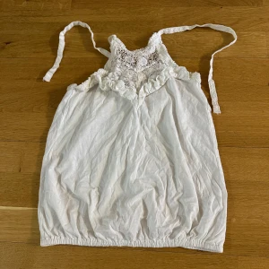 Vit spetsdetaljerad linnetopp - Säljer en söt vit linnetopp med spets och virkade detaljer upptill samt smala axelband som knyts. Storlek XS och köpt utomlands. Knappt använd! Toppen har en luftig passform och resår nertill för en bekväm känsla. Perfekt för varma dagar.