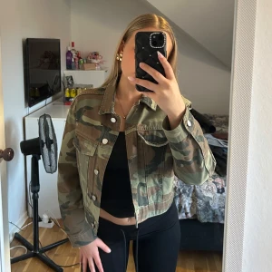 Camo jeansjacka - Säljer en camouflagemönstrad jeansjacka från Gina Tricot i storlek S. Jackan har klassisk krage, knäppning framtill och två bröstfickor med lock. Modellen är croppad och har långa ärmar. 