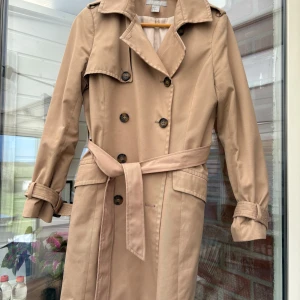 Beige trenchcoat med bälte - Säljer en klassisk beige trenchcoat från H&M med dubbelknäppning och bälte i midjan. Jackan har långa ärmar med knäppning vid ärmslut och bred krage. Perfekt för övergångssäsonger och ger en stilren look.