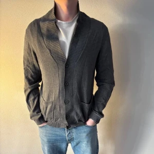 Mörkgrå kofta med krage - Mörkgrå kofta med bred ribbad sjalkrage och knäppning framtill. Två fickor framtill och ribbade muddar vid ärmslut och nederkant. Perfekt för lager-på-lager och enkel att matcha.