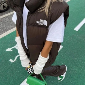 Brun dunväst från The North Face - Säljer en brun dunväst från The North Face med hög krage och dragkedja framtill. Västen har två fickor med dragkedja och logga på bröstet. Perfekt att slänga över en hoodie eller t-shirt för en cool och avslappnad look.