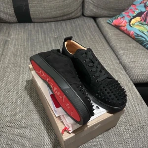 Svarta, Christian Louboutin (bara byta) - Säljer ett par svarta sneakers från Christian Louboutin med ikoniska röda sulor och svarta nitar på tån. Skorna har snörning och är tillverkade i mocka med detaljer i skinn. Perfekta för dig som vill sticka ut med en lyxig och edgy stil.