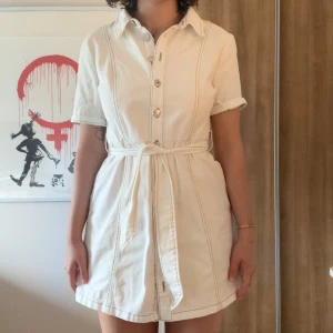 Vit kortklänning med knappar och bälte - Säljer en vit kortklänning med krage, knappar framtill och tillhörande bälte i midjan. Klänningen har korta ärmar och markerade sömmar för en snygg look. Perfekt för varma dagar och enkel att styla.