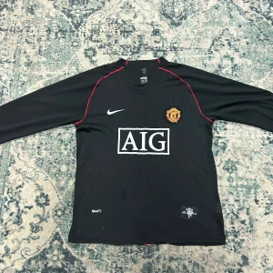 Manchester United långärmad fotbollströja - Säljer en svart långärmad Manchester United-tröja med AIG som sponsor, Nike-logga och klubbmärke på bröstet. På ryggen står det Beckham och nummer 7. Röda detaljer längs axlarna och en röd rand på ryggen. Perfekt för fans av The Red Devils.