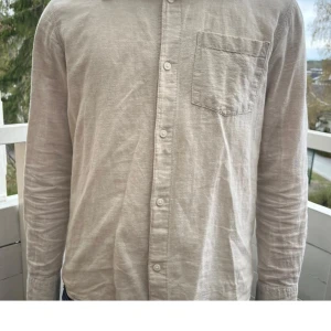 Beige linneskjorta med bröstficka - Säljer en stilren beige skjorta i linne med klassisk krage och bröstficka. Skjortan har långa ärmar och knäpps framtill med knappar. Perfekt för en avslappnad men ändå uppklädd look.