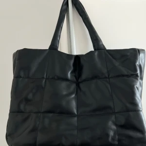Svart quiltad shoppingväska - Stor svart shoppingväska med quiltad design och två handtag. Perfekt för att bära med sig allt man behöver till skolan eller stan. Väskan har en stilren look och är tillverkad i ett material som liknar skinn.