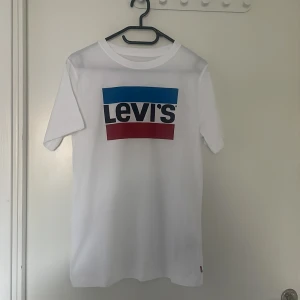 Vit t-shirt med Levi's-logga - Vit t-shirt från Levi's med klassisk stor logga framtill i blått, svart och rött. T-shirten har rund halsringning och korta ärmar. Helt oanvänd och har storleken S. Priset kan diskuteras.