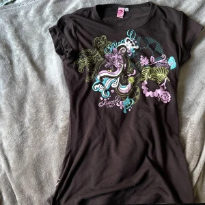 Svart t-shirt med färgglatt tryck från Rip Curl - Brun t-shirt från Rip Curl med färgglatt grafiskt tryck i lila, blått och grönt framtill. T-shirten har rund halsringning och korta ärmar. Materialet känns mjukt och följsamt, perfekt för en avslappnad stil. Även sjukt Y2k. Det står L men det är en extremt liten L (Antagligen för att den är från 2000s talet) Jag skulle säga att det är en S!