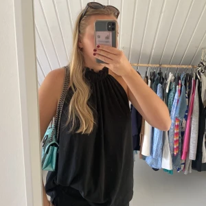 Svart halterneck blus med rynkad krage - Säljer en svart blus med halterneck och rynkad hög krage. Blusen är ärmlös och har en luftig, lös passform. Perfekt för varma dagar eller att styla med kavaj. Materialet känns lätt och faller snyggt.