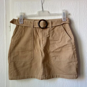Beige kjol med bälte och fickor - Säljer en beige kortkjol med markerad midja och tillhörande bälte med rund spänne. Kjolen har två stora fickor fram och två bak, samt synliga vita sömmar som ger en cool look. Perfekt att styla med en enkel topp eller hoodie.