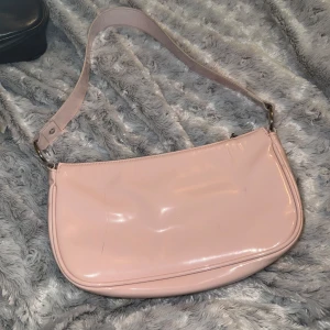 Ljusrosa axelväska i syntet - Säljer en ljusrosa axelväska i glansigt syntetmaterial. Väskan har en enkel, avlång form och stängs med dragkedja upptill. Perfekt för att bära det viktigaste och har en bred axelrem för bekväm användning.