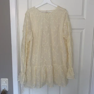 Ljusgul klänning med broderade detaljer från NA-KD - Supersöt ljusgul klänning från NA-KD med broderade blommor, volang nertill och långa ärmar. Klänningen har djup rygg med knytband och en luftig passform. Perfekt till sommaren! Köpte den för 600kr men väljer att sälja för 500kr