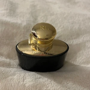 Parfymflaska Ralph Lauren med gulddetaljer - Elegant parfymflaska från Ralph Lauren med lyxig guldfärgad kork och detaljer. Flaskan är svart och ovalformad med en glänsande guldfärgad topp där RL är ingraverat. Perfekt för dig som gillar exklusiva dofter och snygg design.