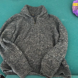 Gråmelerad stickad kofta från Polo Ralph Lauren - Mysig gråmelerad stickad kofta från Polo Ralph Lauren med dragkedja framtill och hög krage. Perfekt för kyliga dagar och enkel att matcha med olika outfits.