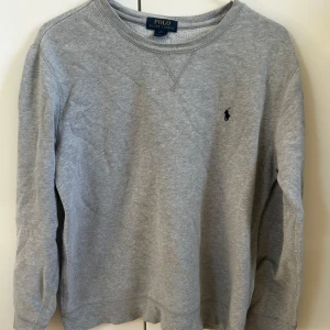 Ralph lauren tröja - Klassisk grå sweatshirt från Polo Ralph Lauren med rund halsringning och broderad logga på bröstet. Tröjan har långa ärmar och är tillverkad i mjuk bomull, perfekt för en avslappnad stil. Köpt på kidsbrandstord för 1199 kr, säljs för endast 299 kr. Tröjan är i nyskick, inga defekter. Hör av dig för fler frågor eller bilder! 🤝 (storlek 14-16 år, motsvarar xs-s. Passar mig som bär S)