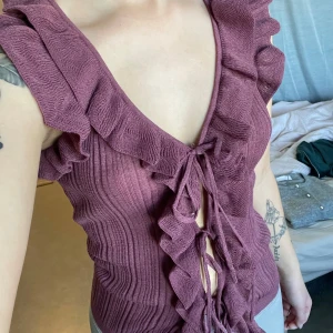 Lila viral Zara topp  - Trendig plommonlila topp från Zara med djup v-ringning, volangdetaljer längs kanterna och snörning framtill. Ärmlös och ribbad för en snygg passform. Perfekt för att lyfta din outfit med en unik touch. Ej använd. Säljer den då jag inte tycker den passade mig. 