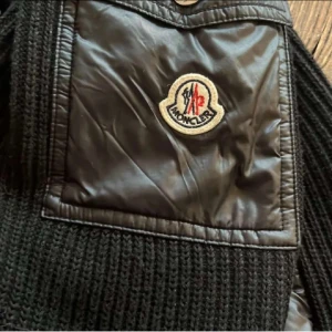 Svart pufferjacka från Moncler - Snygg svart pufferjacka från Moncler med stickade ärmar och vadderad kropp. Jackan har dragkedja framtill, huva och en ficka med Moncler-logga på ärmen. Perfekt för kyliga dagar och riktigt trendig look.