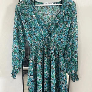 Blommig klänning med v-ringning från Zara - Säljer en blommig klänning från Zara i gröna och blå toner. Klänningen har v-ringning, långa ärmar med resår vid handlederna och smock i midjan. Perfekt för vår och sommar!