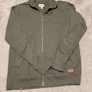 Blend ziptröja - Säljer en mörkgrön ziptröja från blend med hel dragkedja framtill. Tröjan har ribbade muddar vid ärmslut och nederkant samt diskret logodetalj vid höften. Perfekt för lager-på-lager och enkel att matcha.