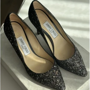 Svarta glittriga pumps från Jimmy Choo - Säljer ett par svarta pumps från Jimmy Choo med glittrig yta och spetsig tå. Skorna har hög klack och är perfekta för att sticka ut. Klassisk och elegant design med lyxig känsla.