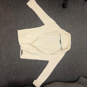 Vit stickad kofta från WILVORST LLOYD - Säljer en stilren vit stickad kofta från WILVORST LLOYD. Koftan har långa ärmar, knappar framtill och ett diskret mönster i stickningen. Perfekt för lager-på-lager och passar till många olika outfits. Väldigt Grisch!