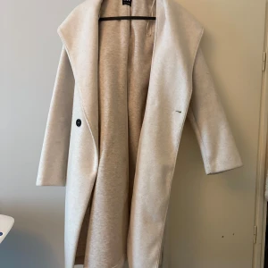 Beige kappa med huva och bälte - Säljer en stilren beige kappa med stor huva och bälte i midjan. Jackan har lång ärm, bred krage och knäpps med en knapp framtill. Perfekt för dig som gillar en klassisk och enkel look.