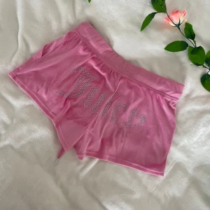 Rosa velourshorts från Juicy Couture - Säljer ett par ikoniska rosa velourshorts från Juicy Couture med glittrande stenar som bildar texten 'Juicy' på baksidan. Shortsen har bred resår i midjan, snörning och fickor fram. Har ett hål på sidan från att jag råkade dra bort prislappen lite för hårt, men det syns inte (sista bilden), annars helt oanvända