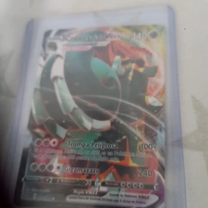 Copperajah VMAX Pokémonkort - Säljer ett Copperajah VMAX Pokémonkort med holografisk yta. Kortet har färger i grönt, orange och silver och är på spanska. Det är inplastat i ett skyddsfodral. Perfekt för samlare eller Pokémonfans!