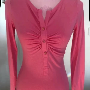 Rosa långärmad topp från Ilse Jacobsen - Säljer en rosa långärmad topp från Ilse Jacobsen. Toppen har knappar framtill, rynkade detaljer vid bysten och en v-ringad hals. Perfekt för dig som gillar en feminin och figurnära stil.