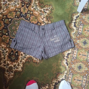 Rutiga shorts med broderi - Snygga gråa rutiga shorts med broderad text på ena sidan. Shortsen har knappar på sidorna och fickor både fram och bak. Perfekta för en trendig look!
