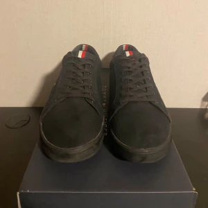 Svarta sneakers från Tommy Hilfiger - Säljer ett par svarta sneakers från Tommy Hilfiger med klassisk låg profil och rund tå. Skorna har snörning och små Hilfiger-detaljer i rött, vitt och blått på plösen och sidan. Perfekta för en stilren och enkel look.