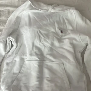 Vit hoodie från GANT med logga - Snygg vit hoodie från GANT med broderad logga på bröstet. Klassisk känguruficka framtill och huva. Mjuk bomullskänsla och stilren design som passar perfekt till jeans eller mjukisbyxor.