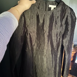 Svart glansig långärmad klänning H&M - En svart långärmad klänning från H&M med glansig, skimrande yta och lätt struktur i tyget. Klänningen har rund halsringning och dragkedja bak. Perfekt för dig som vill sticka ut med en elegant och modern look.
