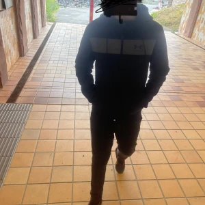 Svart vindjacka Under Armour - Svart vindjacka från Under Armour med grått band över bröstet och logga. Jackan har huva med snörning, dragkedja framtill och långa ärmar. Perfekt för dig som vill ha en sportig och clean look. Storlek: xs