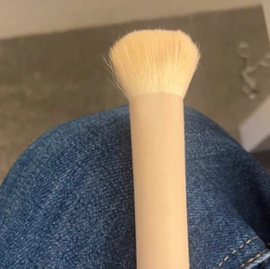 Beige sminkborste med täta strån - En sminkborste med tätt packade, ljusa borst och ett beige handtag. Perfekt för att applicera foundation, bronzer eller puder. Borsten har en platt, rundad form som gör det enkelt att blenda ut produkterna jämnt över huden.